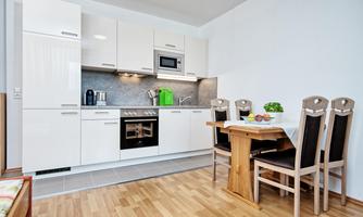 A modern kitchen with white furniture and wooden flooring. A dining table with chairs is next to the kitchen unit.