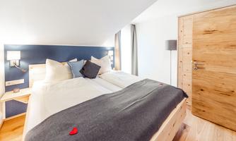 A modern bedroom with a double bed and light wood. The walls are painted blue, and there are cozy cushions as well as a wooden wardrobe door.