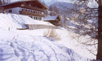A snowy landscape with traditional chalets and mountains in the background. The snow covers the ground, and footprints are visible.