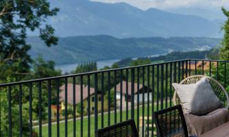 A beautiful view of mountains and a lake from a terrace with a large table and chairs. The surroundings are green and peaceful.