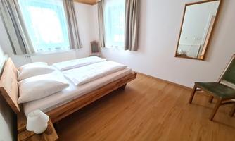 A bright bedroom with a double bed and wooden flooring. The room has large windows with curtains and a mirror on the wall.