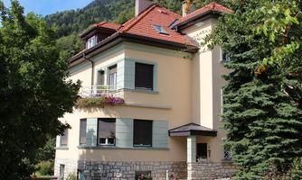 A beautiful house with a red tiled roof and large windows. Surrounded by trees and bushes in a green landscape.