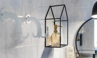 An elegant soap dispenser in a modern, house-like holder. In the background, white hooks and a round mirror are visible.