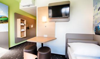 A modern hotel room with a table, two chairs, and a television on the wall. The walls are designed in light colors, and there are shelves with towels.