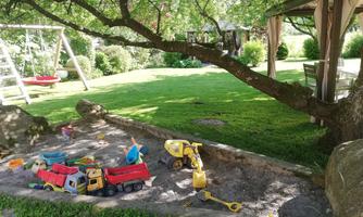 A play area in the garden with a sandpit and colorful toys. In the background, a swing and plenty of greenery can be seen.