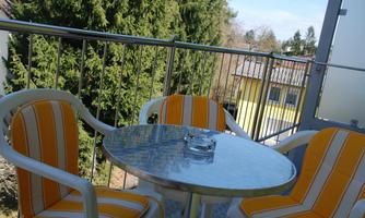 A balcony with two chairs and a round table. The table has a glass top and is positioned among green trees on sunny days.
