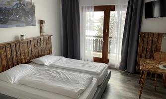 A bright bedroom with two single beds and a rustic wooden headboard. Outside is a balcony with a view and a cozy sitting area in the room.