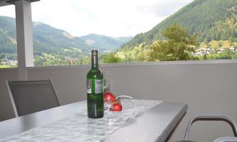 A table on a balcony with a green bottle and apples. In the background, there are wooded mountains and a clear sky.