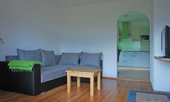 A simple living room with a gray couch and a wooden table. In the background, an open kitchen is visible.
