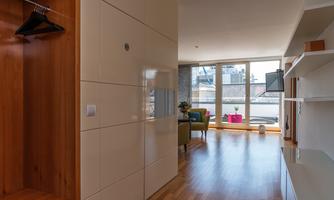 A modernly furnished room with wooden flooring and large windows that provide plenty of daylight. In the background, a cozy seating area and a television can be seen.