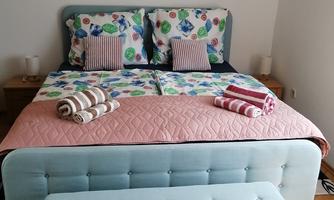 A cozy bedroom with a large double bed and colorful pillows. Freshly rolled towels are lying on the bed, and an elegant stool stands at the foot of the bed.