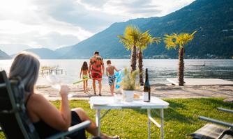 A family is walking by the shore of a lake, while a woman is relaxing on a lounge chair. In the background, palm trees and mountains can be seen.