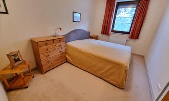 A simple bedroom with a large bed and a wooden dresser. The room has a pleasant, neutral tone and a window with curtains.