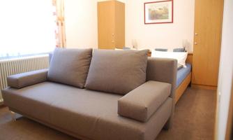 A simply designed room with a beige sofa and a double bed. The walls are light, and there is a small table as well as a wardrobe in the background.