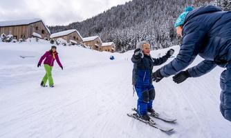 A child is learning to ski in a snowy environment. In the background, wooden huts and snow-covered mountains can be seen.