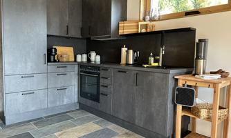A modern kitchen with dark cabinets and a practical wooden shelf. The windows let in a lot of daylight.