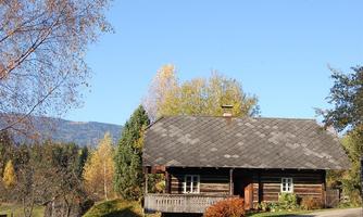 A charming wooden house surrounded by trees and colorful autumn leaves. In the background, gentle hills and a clear blue sky are visible.
