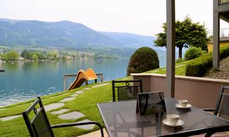 A beautiful view of a tranquil lake with green hills in the background.  
On the shore, there is a table with cups and a sliding area for children.