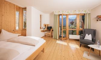 A cozy hotel room with wooden furniture and large windows. The room offers a view of the mountains and has a desk as well as a comfortable armchair.