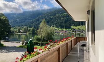 A cozy terrace with a view of a tranquil lake and green mountains. Colorful flowers and chairs invite you to relax.