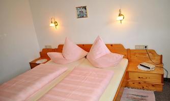A cozy bedroom with two single beds and pink cushions. Two wall lamps and a wooden dresser complete the decor.