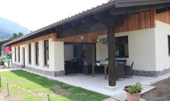 A modern house with wooden cladding and a large terrace. The garden is well-maintained and surrounded by mountains.