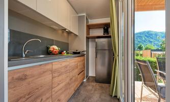 A modern kitchen with wood fronts and a stainless steel sink. Through the window, a green landscape and a blue sky can be seen.