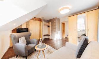 A cozy living room with a gray armchair and a sofa. The space is bright and modernly designed, featuring wood and neutral color tones.