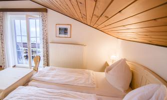 A cozy bedroom with two beds and a wooden ceiling. The room has windows with a view outside and soft lighting.