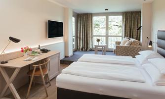 A modern hotel room with two beds, a desk, and a television. Large windows let in plenty of daylight and offer a view of the outdoor area.