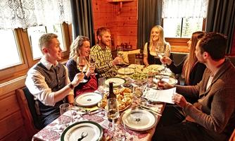 A group of people is sitting at a table in a cozy wooden cabin. They are toasting with glasses and enjoying a festive meal.