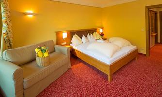 A cozy hotel room with a large bed and a comfortable couch. The walls are painted in a warm yellow tone, and the room is well-lit.