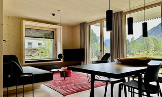 A modern living room with large windows and a view of nature. There is a black couch, a table, and decorative elements like a red carpet.