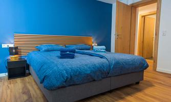 A modern bedroom with a large blue bed and an elegant wooden headboard. The walls are painted blue, and there is a bedside lamp as well as fresh towels.