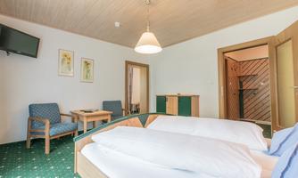 A cozy hotel room with two single beds, a television, and a table. The decor is simple and inviting with green carpet and light walls.