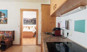 A modern kitchen with wooden cabinets and a cooktop. In the background, a cozy bedroom with a large bed is visible.