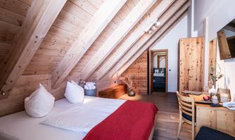 A cozy attic room with wooden walls and a large window.  
There is a bed with white pillows and a small seating area with a table.