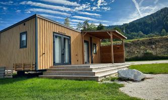 A modern wooden house with a porch is surrounded by green grass. In the background, gentle hills and a blue sky with clouds can be seen.