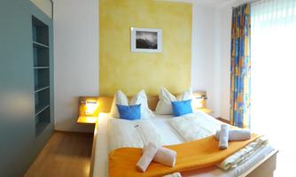 A cozy bedroom with a double bed and colorful cushions. The walls are painted in warm colors, and daylight streams through the windows.