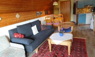 A cozy living room with a black sofa, a wooden table, and a rug. In the background, there is a small kitchen and a dining area with chairs.