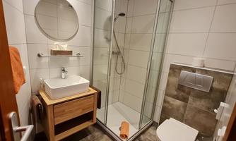 A modern bathroom with a shower and a wooden sink. The walls are tiled in light colors, and there are orange-red towels for decoration.