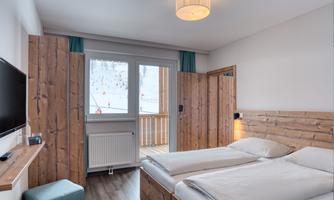 A cozy room with wooden furniture and two single beds. The view is of a balcony from which you can see the mountains.