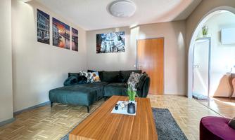 A modern living room with a green sofa and a wooden table. City pictures hang on the wall, and there is an open transition to the kitchen.