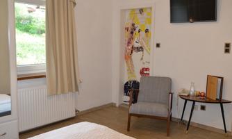 A bright room with a bed and an armchair. A television is hanging on the wall and a colorful art print is visible.