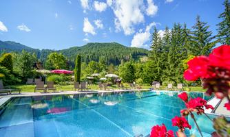 An inviting pool surrounded by green mountains and colorful flowers. In the background, sunny loungers and sun umbrellas can be seen.