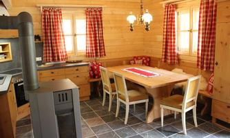 An inviting dining area in a wooden cabin with a large wooden table and six white chairs. The walls are made of wood and the windows have red checkered fabric curtains.