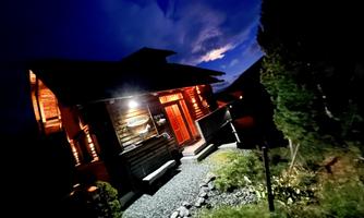 A charming wooden house at night, illuminated by warm light. Surrounded by green plants and a clear sky.