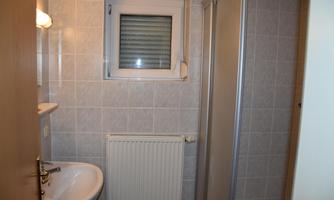 A small bathroom with a shower, a sink, and a window. The walls are covered with light tiles.