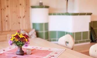 A cozy table with a flower vase and a red tablecloth. In the background, a traditional tiled stove can be seen.