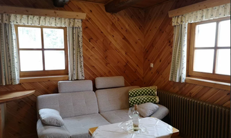 A cozy room with wooden paneling and large windows. A gray sofa and a table with a bottle and a tablecloth are also visible.